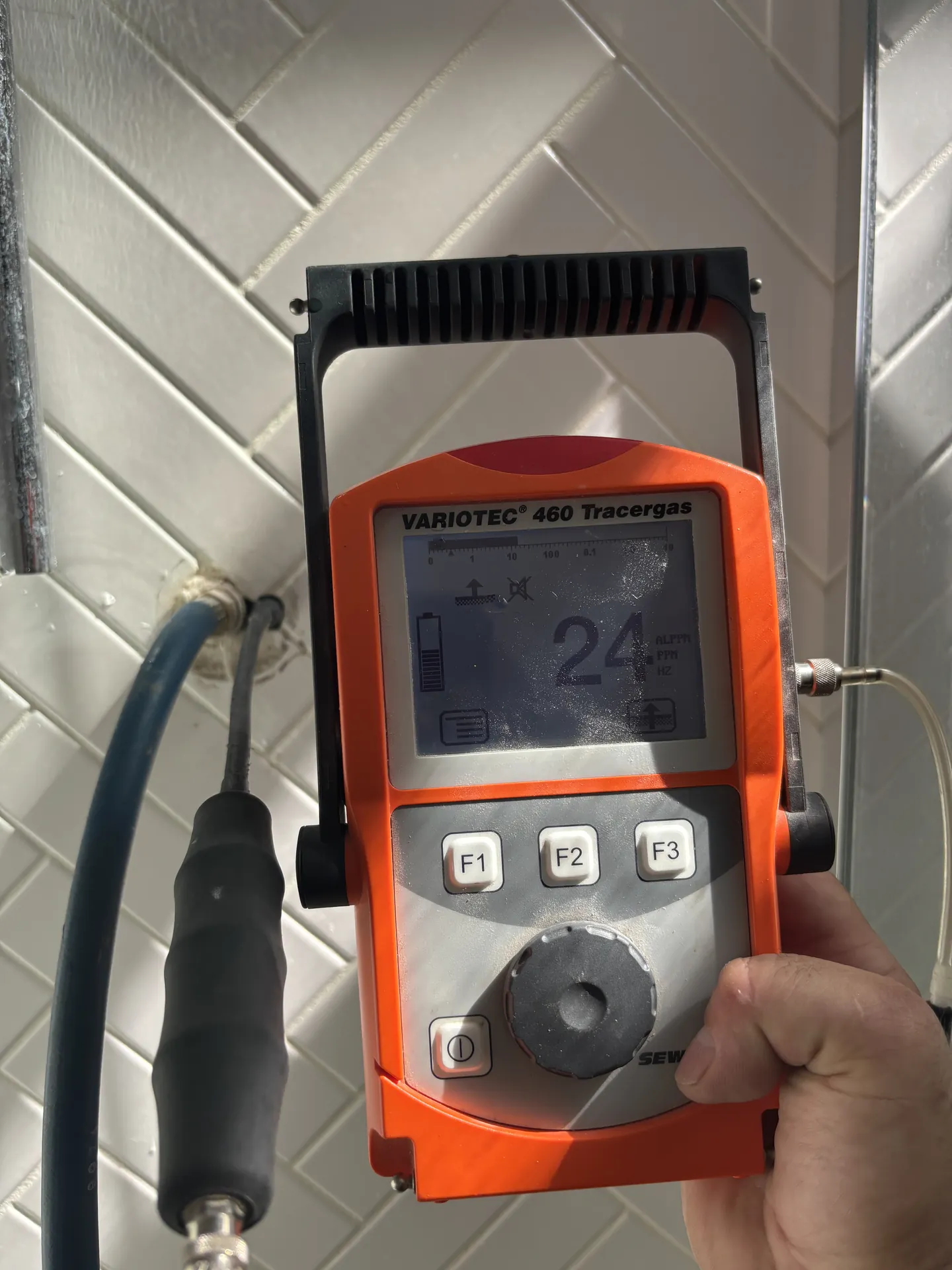 Tracer gas detector reading 24 at a herringbone-tiled bathroom wall